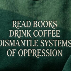 Op de afbeelding: Een donker bosgroen sweatshirt met witte geborduurde tekst die luidt: READ BOOKS DRINK COFFEE DISMANTLE SYSTEMS OF OPPRESSION. De stof heeft een zachte, fleece-achtige textuur en de tekst is gecentreerd in een strak, schreefloos lettertype.