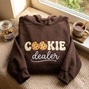 Può includere: Una felpa con cappuccio marrone scuro con testo ricamato che recita "COOKIE dealer" in un carattere color crema. Le due lettere O nella parola cookie sono sostituite da illustrazioni di biscotti con gocce di cioccolato e una stella rossa.