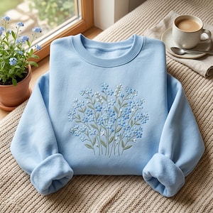 May include: A light blue crewneck sweatshirt featuring a central embroidered design of a bouquet of small blue forget-me-not flowers with green stems and leaves. The garment has long sleeves with ribbed cuffs and a ribbed neckline, showcasing detailed stitching.