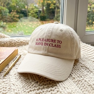 May include: A cream-coloured baseball cap featuring the embroidered text A PLEASURE TO HAVE IN CLASS in dark red lettering. The cap has a structured crown, a curved brim, and stitched eyelets on the top panels for ventilation and durability.