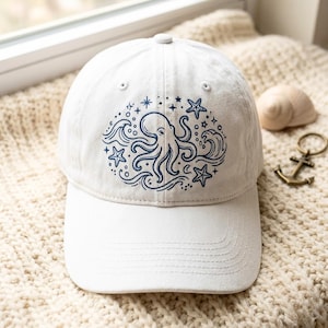 Op de afbeelding: Een witte baseballpet met een donkerblauwe, cirkelvormige grafische print op de voorzijde. Het ontwerp toont een octopus in het midden, omringd door gestileerde oceaangolven, kleine sterren en zeesterren in een strakke lijntekeningstijl.