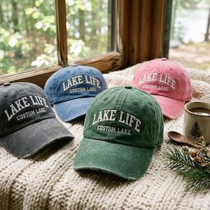 Könnte beinhalten: Drei Baseballkappen in Anthrazit, Blau und Grün sowie eine rosafarbene Kappe, alle mit weißem Sticktext LAKE LIFE und CUSTOM LAKE. Jede Kappe hat einen abgenutzten Schirm und eine strukturierte Krone mit Ösen für die Belüftung.