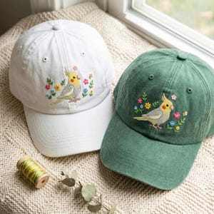 Peut inclure: Une casquette de baseball blanche et une casquette de baseball vert foncé, chacune ornée d'une broderie représentant une calopsitte grise et jaune entourée de petites fleurs colorées. Une bobine de fil jaune est posée à proximité.
