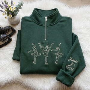 Puede incluir: Una sudadera verde oscuro con cremallera de cuarto de cuello que presenta un bordado de líneas blancas de tres patinadores artísticos en movimiento. La manga derecha tiene bordado el nombre Janie en cursiva sobre un gráfico de patín.