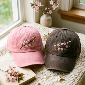 Puede incluir: Dos gorras de béisbol, una en rosa desteñido y otra en gris carbón oscuro, cada una con una rama bordada de flores de cerezo de color rosa claro. Las gorras tienen una corona estructurada, una visera curva y ojales para ventilación.