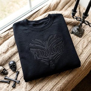 Peut inclure: Un sweat-shirt noir à col rond avec une broderie ton sur ton sur la poitrine. La broderie représente une cage thoracique humaine en forme de cœur avec le mot RADIOLOGY intégré au centre de la structure squelettique.