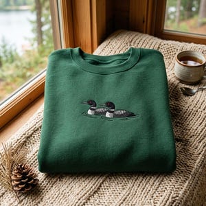 Peut inclure: Un sweat-shirt à col rond vert forêt orné d'une broderie représentant deux plongeons nageant sur l'eau. Les oiseaux sont représentés avec des têtes noires, un dos tacheté de blanc et des yeux rouges, centrés sur la poitrine du vêtement à manches longues.
