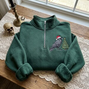 Könnte beinhalten: Ein dunkelgrüner Sweatshirt mit Viertelreißverschluss und einer Stickerei auf der Brust. Die Stickerei zeigt eine schwarze Krähe mit einer rot-weißen Weihnachtsmannmütze, die neben einem geschmückten grünen Weihnachtsbaum mit kleinen gelben Lichtern steht.