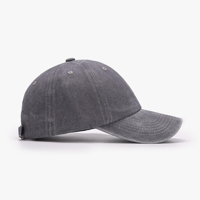May include: A charcoal gray baseball cap with a washed, distressed fabric finish. The cap features a six-panel construction with stitched eyelets, a curved brim with horizontal stitching, and a small fabric-covered button at the crown center.