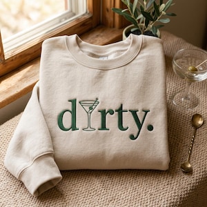 Op de afbeelding: Een beige sweater met ronde hals en donkergroene geborduurde tekst "dirty.", waarbij de letter "i" is vervangen door een afbeelding van een martiniglas. Het kledingstuk heeft lange mouwen, een geribbelde halslijn en een zachte, effen stof.