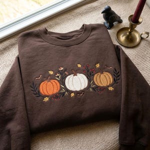 May include: A dark brown crewneck sweatshirt featuring an embroidered design across the chest. The embroidery depicts three pumpkins in orange, white, and tan, surrounded by dark green foliage, small red berries, yellow leaves, and tiny black bats.