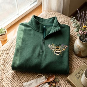 May include: A dark green quarter-zip pullover featuring an embroidered bee on the left chest. The bee has yellow and black stripes on its body and wings decorated with small, colorful floral patterns in shades of pink, purple, and yellow.