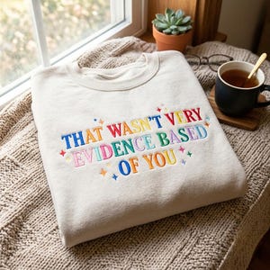 Könnte beinhalten: Ein beigefarbenes Sweatshirt mit Rundhalsausschnitt und mehrfarbigem Sticktext "THAT WASN'T VERY EVIDENCE BASED OF YOU", verziert mit kleinen Sternen in Blau, Rot, Gelb, Grün und Pink, die sich über den Brustbereich des Kleidungsstücks erstrecken.