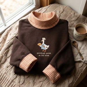 May include: A dark brown long-sleeve sweatshirt with a mock neck collar and sleeve cuffs featuring an orange and white gingham pattern. The center chest displays an embroidered goose wearing a chef hat and apron, holding bread, with the text "sourdough goose on the loose" embroidered below the graphic.