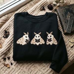 May include: A black crewneck sweatshirt featuring an embroidered design of three fluffy cream-colored moths, each holding a small open book. The moths have detailed brown wing patterns, large dark eyes, and feathery antennae, arranged horizontally.