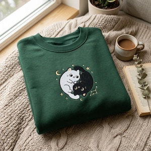 Könnte beinhalten: Ein dunkelgrüner Rundhals-Pullover mit einer Stickerei, die eine weiße und eine schwarze Katze zeigt, welche sich in einer kreisförmigen Yin-Yang-Form aneinander schmiegen, umgeben von kleinen goldenen Sternen, einer Mondsichel und einem Sternbild.