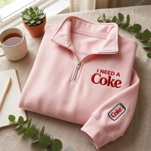 以下が含まれることがあります： 胸元に赤い刺繍で「I NEED A Coke」の文字が施され、左袖には「Coke」と書かれたソーダ缶の小さな刺繍があしらわれた、ライトピンクのクォータージッププルオーバースウェットシャツ。シルバーカラーの金属製ジッパーが付いています。