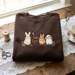 Op de afbeelding: Een donkerbruine sweater met ronde hals voorzien van een borduursel van vier konijnen in diverse tinten crème, bruin en wit, op een rij geplaatst met kleine groene bladaccenten aan de onderkant van het borduurwerk op de voorzijde.