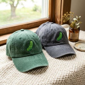 May include: Two baseball caps, one in a muted green and one in a charcoal gray, each featuring an embroidered green pickle graphic on the front panel. Both caps have a structured crown, a curved brim, and a washed, vintage-style fabric texture.
