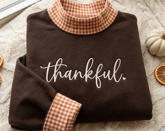 May include: A dark brown long-sleeve sweatshirt featuring a mock neck collar and sleeve cuffs in an orange and white gingham pattern. The word thankful is embroidered in white cursive script across the centre chest, followed by a small heart.