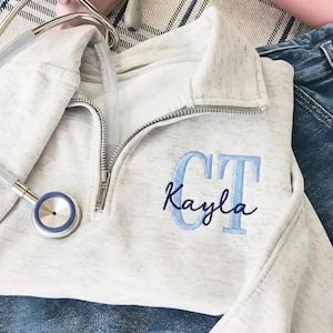Op de afbeelding: Een gemêleerde off-white trui met een korte rits, een kraag en een zilverkleurige metalen rits. Op de borst zijn de initialen CT geborduurd in lichtblauwe blokletters, met de naam Kayla in donkerblauw cursief schrift eroverheen gestikt.