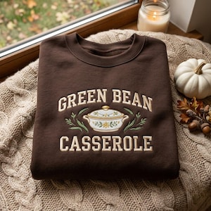 May include: A dark brown crewneck sweatshirt featuring an embroidered design of a white casserole dish with floral accents, flanked by green leaves. The text "GREEN BEAN CASSEROLE" is embroidered in cream-colored block letters above and below the dish.