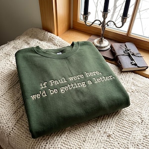 Puede incluir: Una sudadera de cuello redondo en color verde bosque con texto bordado en blanco que dice: "if Paul were here, we'd be getting a letter." La prenda está doblada cuidadosamente, mostrando la costura limpia de las letras sobre la tela oscura.