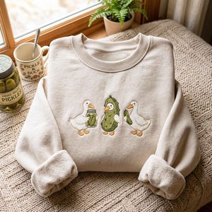 K&ouml;nnte beinhalten: Ein beigefarbenes Sweatshirt mit Rundhalsausschnitt und einer Stickerei von zwei wei&szlig;en Enten sowie einer mittleren Ente, die als Gew&uuml;rzgurke verkleidet ist. Eine Ente h&auml;lt ein Glas mit Gurken, die andere eine einzelne Gurke. Der Stoff ist weich und flauschig.