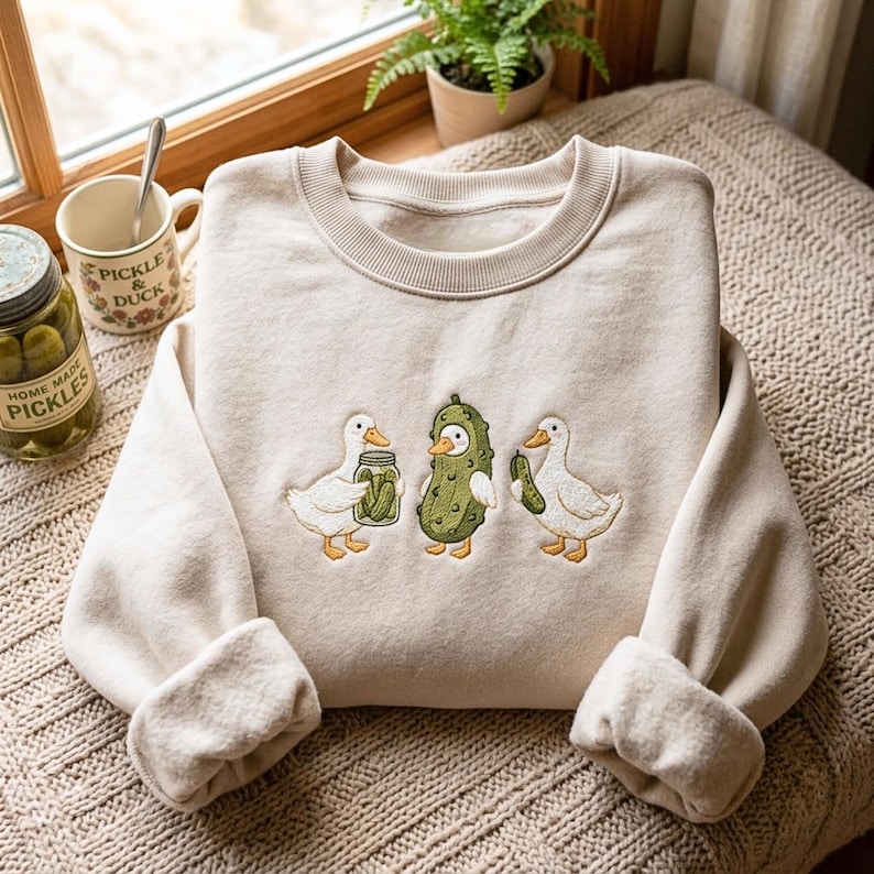K&ouml;nnte beinhalten: Ein beigefarbenes Sweatshirt mit Rundhalsausschnitt und einer Stickerei von zwei wei&szlig;en Enten sowie einer mittleren Ente, die als Gew&uuml;rzgurke verkleidet ist. Eine Ente h&auml;lt ein Glas mit Gurken, die andere eine einzelne Gurke. Der Stoff ist weich und flauschig.