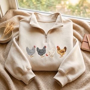 May include: A cream-colored quarter-zip sweatshirt featuring an embroidered design of four chickens in a row. The chickens are depicted in shades of gray, black, white, and brown. The sweatshirt has long sleeves with ribbed cuffs and a high collar.