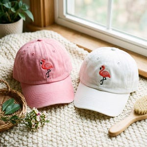 May include: Two baseball caps, one in light pink and one in white, each featuring a small, embroidered pink flamingo graphic on the front panel. Both caps have a curved brim, a button on the top, and stitched eyelets for ventilation.
