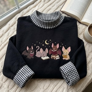 May include: A black long-sleeve sweatshirt featuring a mock neck collar and sleeve cuffs in a black and white gingham pattern. The chest displays an embroidered graphic of four small bats reading books under a yellow crescent moon and stars.