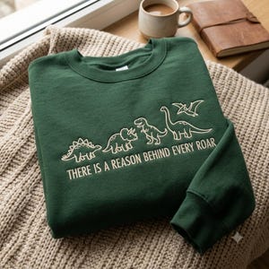Op de afbeelding: Een donkergroene sweater met ronde hals en een witte lijntekening van vier dinosaurussen en een pterosauriër. Onder de dinosaurussen staat de tekst "THERE IS A REASON BEHIND EVERY ROAR" in een strak, schreefloos lettertype op de borst.
