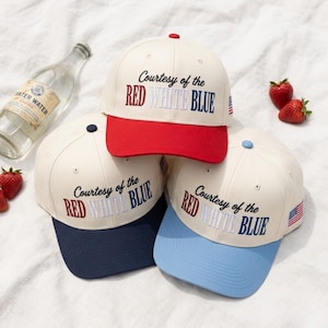 Op de afbeelding: Drie baseballpetten met gebroken witte kronen en geborduurde tekst Courtesy of the RED WHITE BLUE. Eén pet heeft een rode klep, één een marineblauwe klep en één een lichtblauwe klep. Elke pet is voorzien van een kleine Amerikaanse vlag.