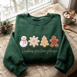 以下が含まれることがあります： ダークグリーンの長袖スウェットシャツ。雪だるま、雪の結晶、クリスマスツリー、ジンジャーブレッドマンのクリスマス・クッキーの刺繍が施されており、その下にゴールドの糸で「Cookies for two please」という文字が刺繍されています。