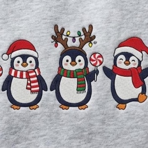 Peut inclure: Un sweat-shirt gris clair avec une broderie repr&eacute;sentant cinq pingouins. Chaque pingouin porte un bonnet de P&egrave;re No&euml;l rouge. Les pingouins tiennent divers objets de f&ecirc;te, notamment une canne en sucre, une &eacute;charpe ray&eacute;e, des bois de renne, une sucette et un cadeau.