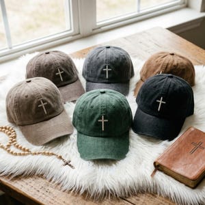 Könnte beinhalten: Eine Sammlung von sechs Baseballkappen in verschiedenen Farben, darunter Hellbraun, Dunkelgrau, Braun, Grün und Schwarz. Jede Kappe hat ein einfaches, weiß gesticktes Kreuz auf der Vorderseite. Der Stoff der Kappen wirkt verwaschen und abgenutzt.