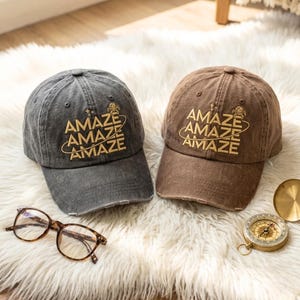 May include: Two baseball caps, one charcoal gray and one brown, each featuring the embroidered text AMAZE stacked three times with a circular orbit line and small star icons. The caps have a distressed brim and are paired with glasses and a compass.