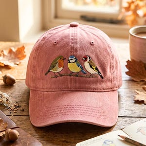 Op de afbeelding: Een oudroze baseballpet met een borduursel van drie kleine vogels op een tak. De vogels zijn een roodborstje met een oranje borst, een pimpelmees met geel en blauw verenkleed, en een putter met een rode kop en zwarte vleugels.