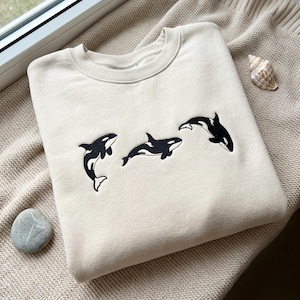 Könnte beinhalten: Ein beigefarbenes Sweatshirt mit Rundhalsausschnitt und einer Stickerei von drei schwarz-weißen Orcas, die in horizontaler Formation in der Mitte der Brust schwimmen. Der Stoff hat eine glatte Textur und eine durchgehend hellbraune Farbe.