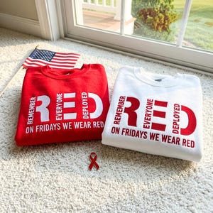 Könnte beinhalten: Ein rotes Sweatshirt und ein weißes Sweatshirt, jeweils mit dem Text REMEMBER EVERYONE DEPLOYED und ON FRIDAYS WE WEAR RED in Kontrastfarben. Eine kleine amerikanische Flagge und eine rote Schleifenanstecknadel liegen bei den beiden Langarmshirts.