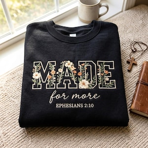 Puede incluir: Una sudadera negra de cuello redondo con la palabra MADE en letras grandes de bloque rellenas con diseños florales bordados en blanco, naranja y verde. Debajo, el texto dice for more y EPHESIANS 2:10 en una fuente blanca cursiva y de bloque.