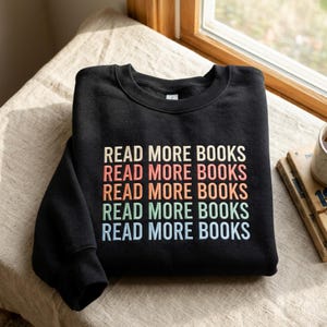 Peut inclure: Un sweat-shirt noir à col rond avec le texte READ MORE BOOKS imprimé cinq fois en lignes superposées. Chaque ligne de texte apparaît dans une couleur différente, notamment crème, corail, orange, vert clair et bleu clair, centrée sur la poitrine.