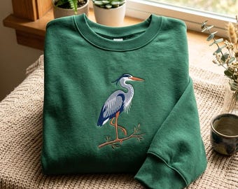 Puede incluir: Una sudadera de cuello redondo en color verde bosque con un diseño bordado de una garza gris y azul posada sobre una rama marrón. El ave tiene un pico largo naranja y patas naranjas, con detalles de plumaje blanco y negro en el cuerpo.