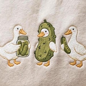 K&ouml;nnte beinhalten: Ein cremefarbener Stoff mit einer Stickerei von zwei wei&szlig;en Enten mit orangefarbenen Schn&auml;beln und F&uuml;&szlig;en. Eine Ente h&auml;lt ein Glas mit gr&uuml;nen Gew&uuml;rzgurken, die andere h&auml;lt eine einzelne Gurke. Dazwischen steht eine Ente in einem Gurkenkost&uuml;m.