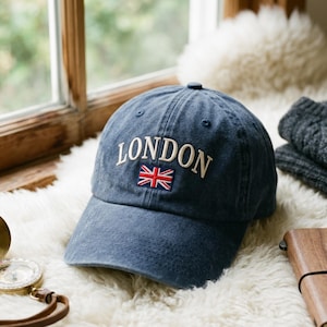 May include: A navy blue baseball cap featuring the word LONDON embroidered in white capital letters above a small, rectangular Union Jack flag patch. The cap has a structured crown, a curved brim, and visible stitching details across the fabric.