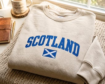 Puede incluir: Una sudadera beige de cuello redondo con la palabra SCOTLAND bordada en letras mayúsculas azules y un icono de la bandera escocesa azul y blanca colocado justo debajo del texto. La prenda tiene puños y cuello acanalados.