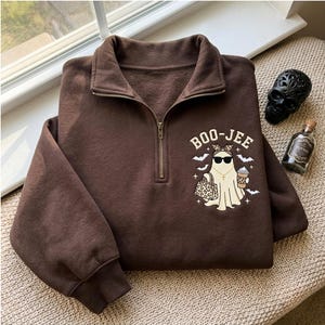 May include: A dark brown quarter-zip sweatshirt featuring a graphic of a ghost wearing sunglasses, a leopard print headband, and a gold necklace. The ghost holds a leopard print handbag and a coffee cup, surrounded by bats and the text BOO-JEE.