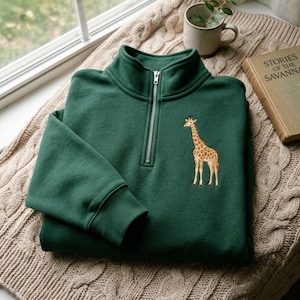 Puede incluir: Una sudadera de color verde oscuro con cremallera de un cuarto, que presenta un cierre metálico plateado en el cuello y un gráfico bordado de una jirafa en el pecho izquierdo. La prenda tiene mangas largas con puños acanalados.