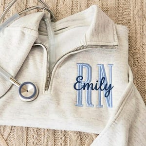 May include: A light heather gray quarter-zip pullover featuring a silver metal zipper and a stethoscope resting on the collar. The chest is embroidered with the light blue letters RN and the name Emily in dark blue cursive script lettering.
